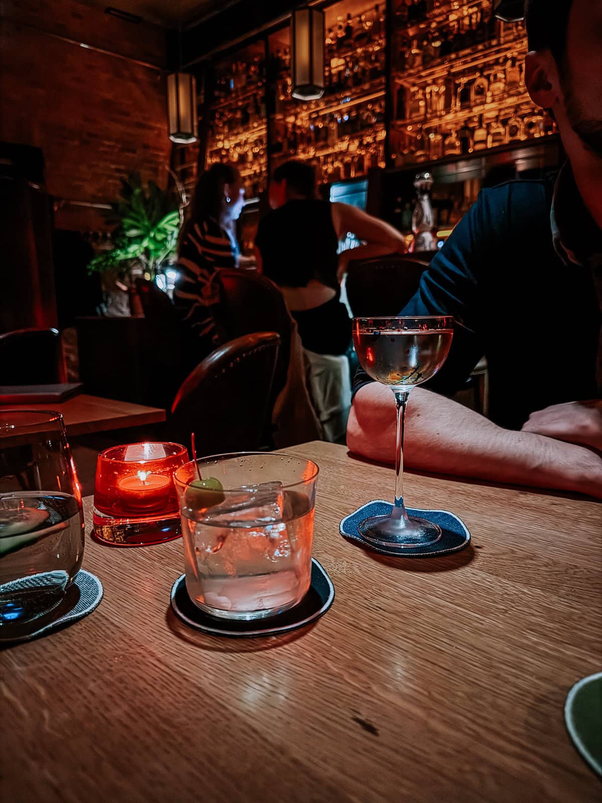 Close-up of drinks on a table in a dimly lit bar, with two people talking in the background near the bar counter