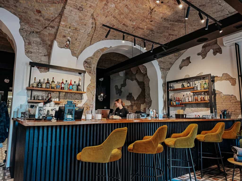 A stylish bar interior featuring a rustic brick ceiling with exposed patches, a wooden bar counter, and several mustard yellow bar stools. The bar shelves are stocked with various bottles and glassware, with a bartender working in the background. The overall ambiance is cozy and inviting, blending modern design with rustic elements.