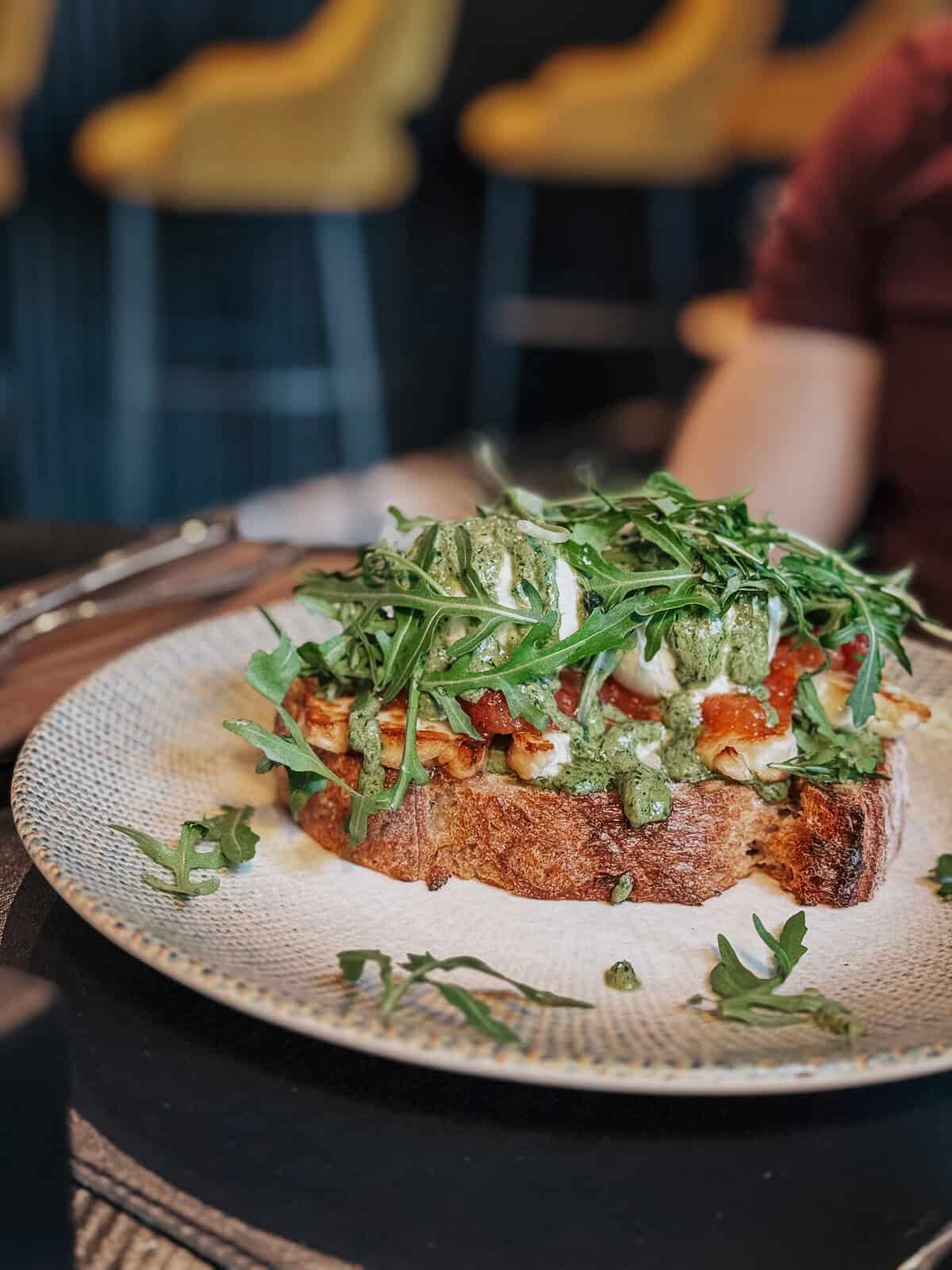 A slice of rustic bread topped with poached eggs, greens, and a drizzle of pesto sauce, set on a patterned plate.