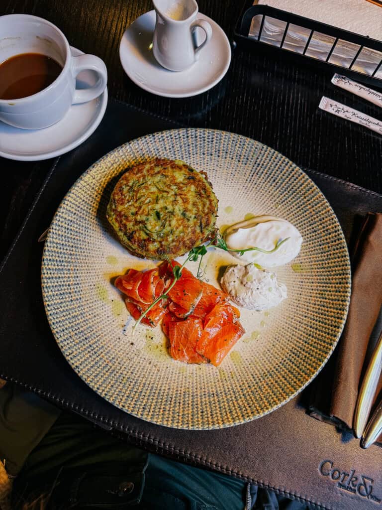 A brunch plate with a potato pancake, smoked salmon, and a dollop of cream cheese, accompanied by a cup of coffee and a small jug of cream.