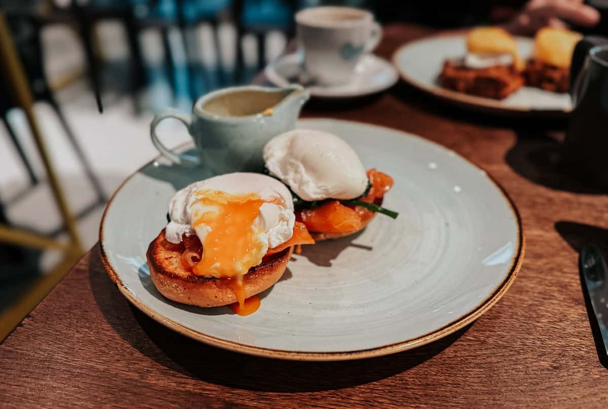 A plate with two poached eggs on toast with smoked salmon and a creamy sauce, with a cup of coffee in the background