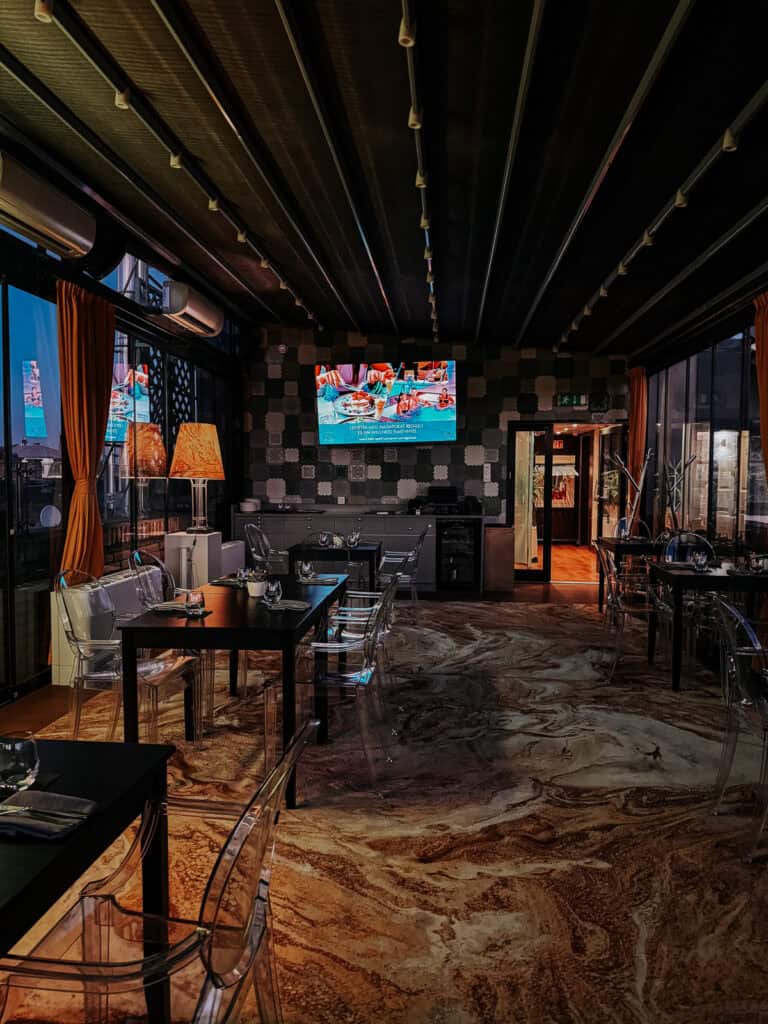 A stylish indoor dining area with contemporary decor, featuring dark tables, transparent chairs, and a TV displaying colorful visuals.