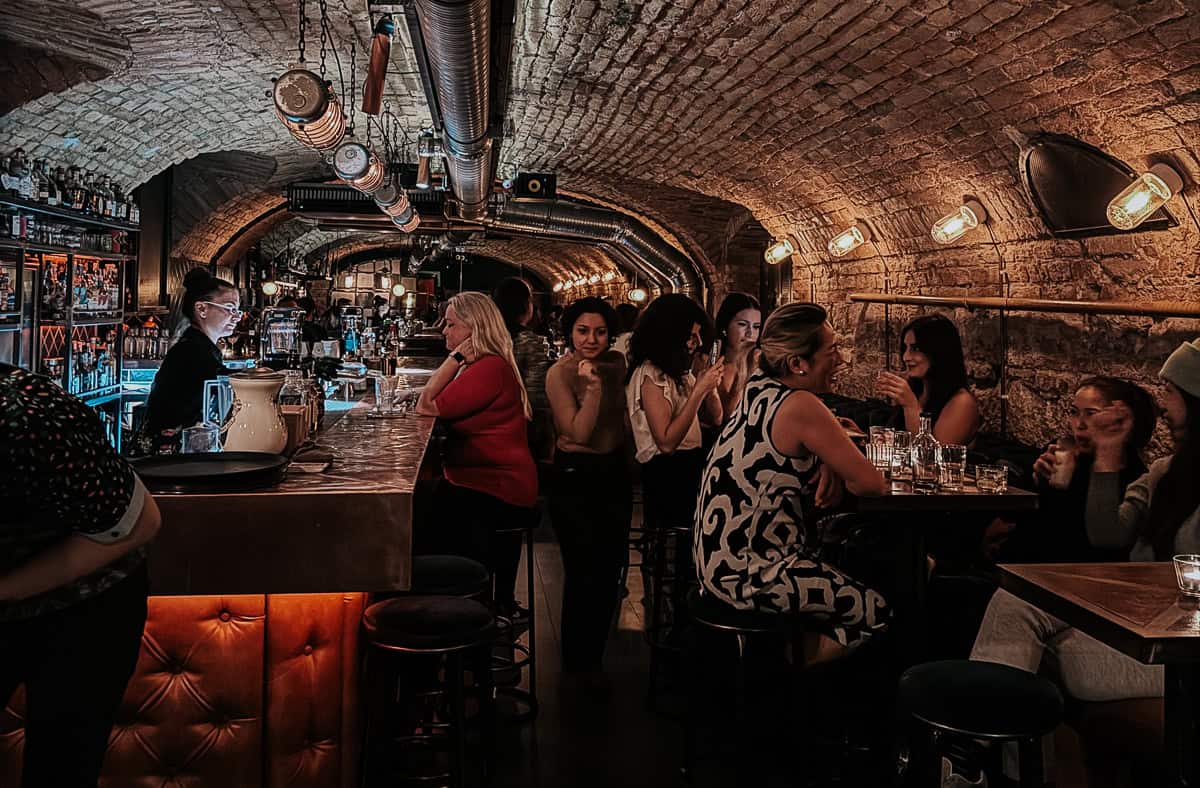 A cozy bar scene with patrons seated at tables and along the bar, featuring an arched brick ceiling and warm lighting.