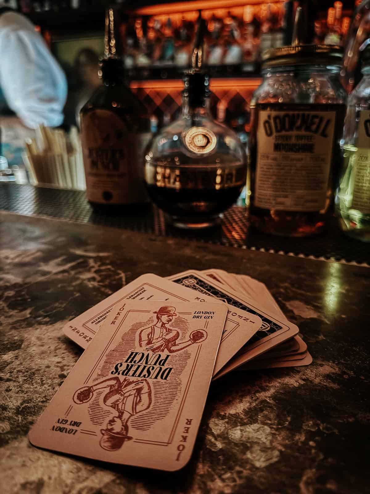 Close-up of a deck of cards and various bottles of spirits on a bar counter, with a warm, inviting glow.