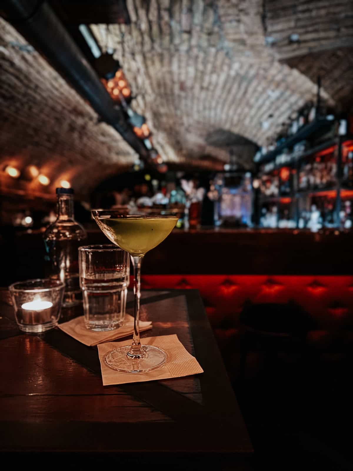 A cocktail served in a martini glass on a small table, with a candle and water glasses, set against a backdrop of an arched brick ceiling and softly lit bar.