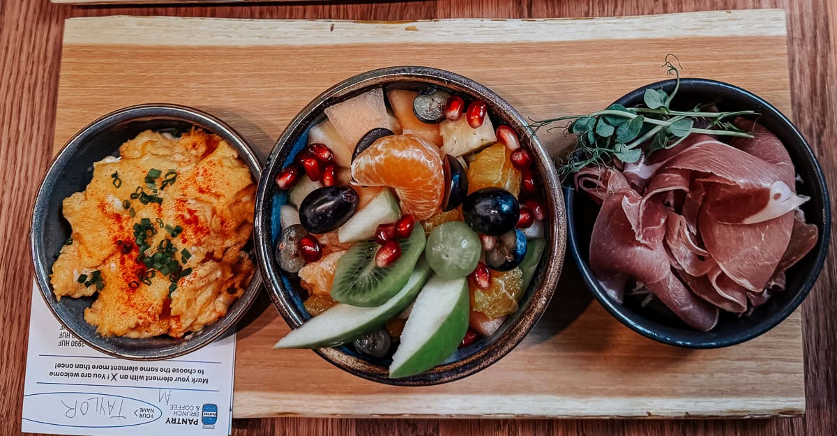 Three small bowls on a wooden board, containing scrambled eggs, a fruit salad with kiwi, grapes, and pomegranate seeds, and a serving of prosciutto.