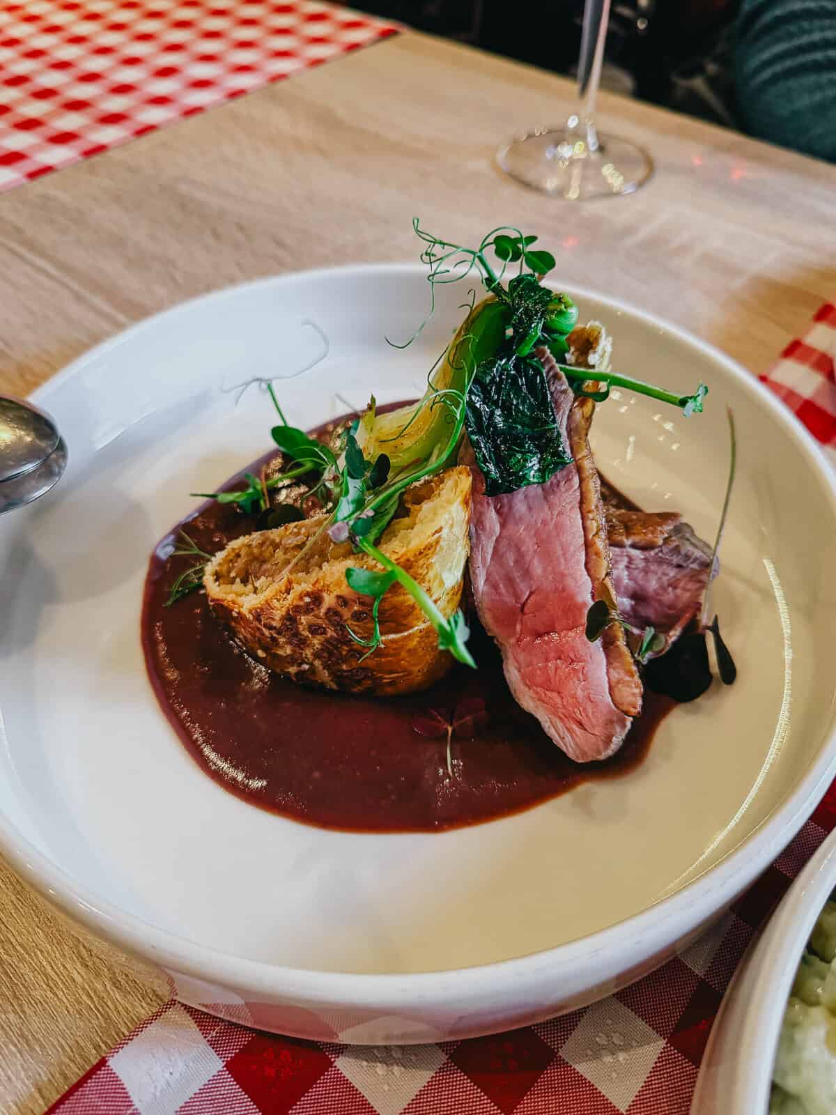 A gourmet dish served in a white bowl, featuring a piece of seared meat, garnished with greens, and accompanied by a sauce.