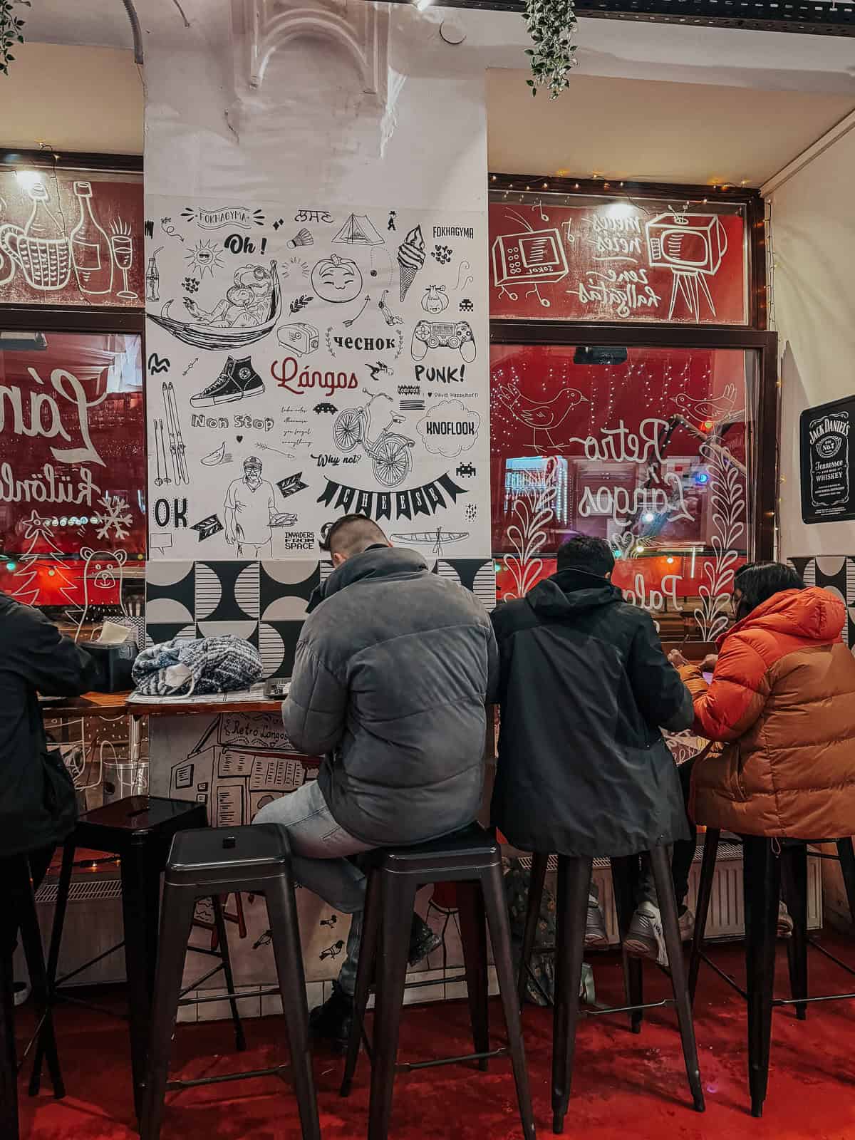 The interior of Retro Lángos, with customers sitting on high stools at a counter. The walls are decorated with whimsical drawings and phrases, creating a lively and nostalgic atmosphere.