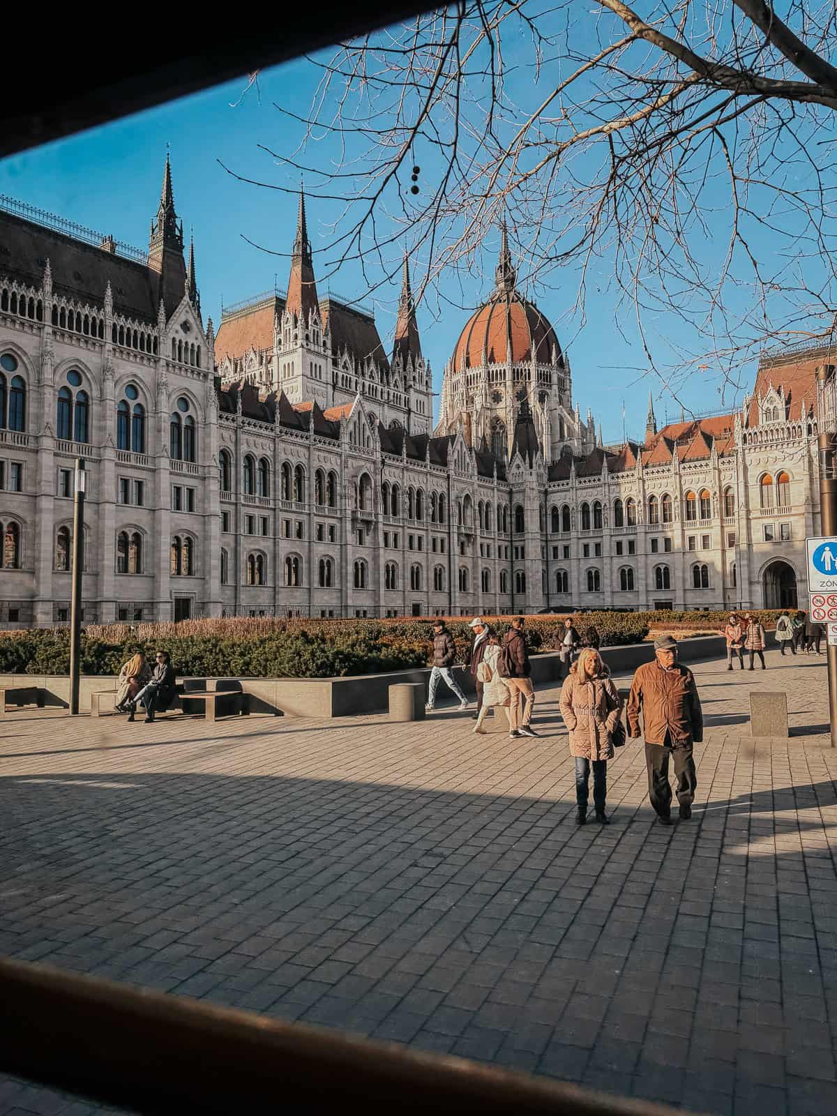 View of the Hungarian Parliament Building in Budapest, with its gothic architecture and red dome, seen from inside a tram. Pedestrians walk and sit in the foreground plaza under a clear blue sky.