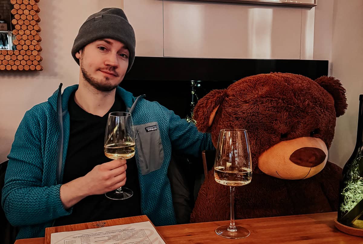 A man in a beanie sitting next to a large teddy bear at a restaurant table, both holding glasses of white wine. The setting is playful and cozy.