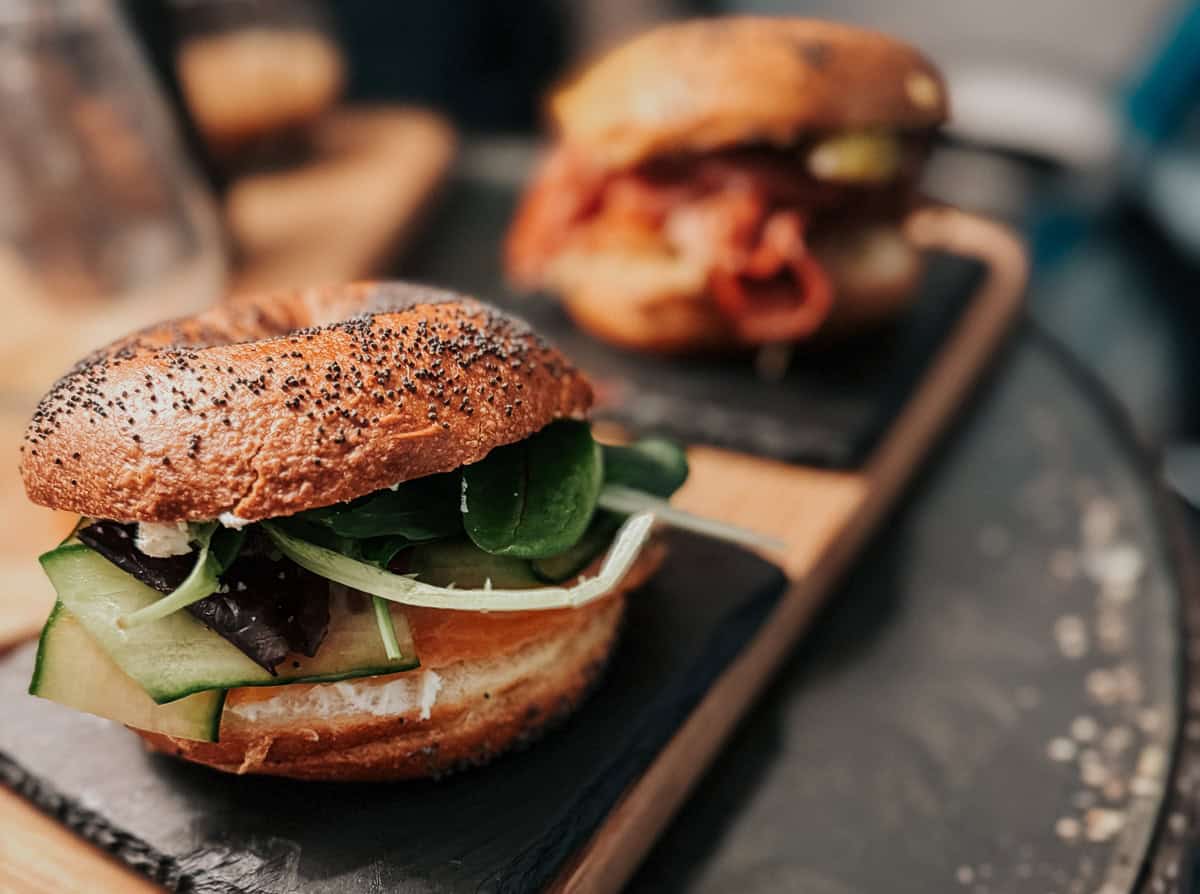 A close-up of a bagel sandwich filled with fresh greens and cucumber slices, with another sandwich in the background.