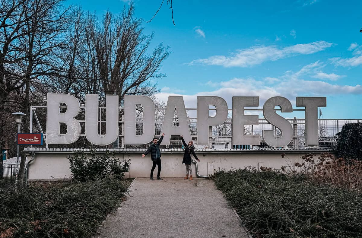 A person sits by the riverbank with Buda Castle in the background in Budapest, Hungary, under a clear sky.