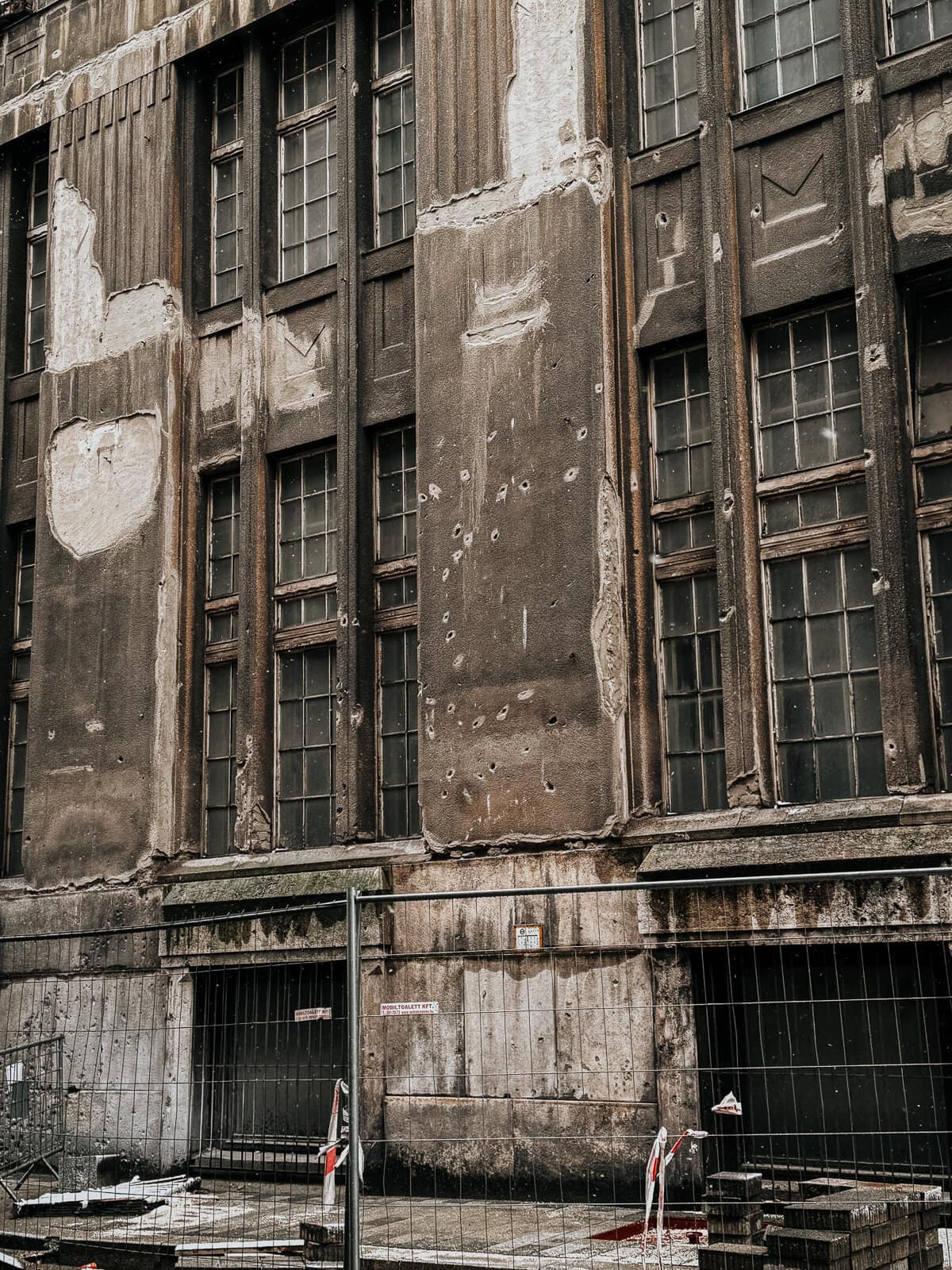 A tall, dilapidated building with numerous bullet holes and patches on its weathered facade. The ground level is blocked off by a metal fence, indicating restricted access.