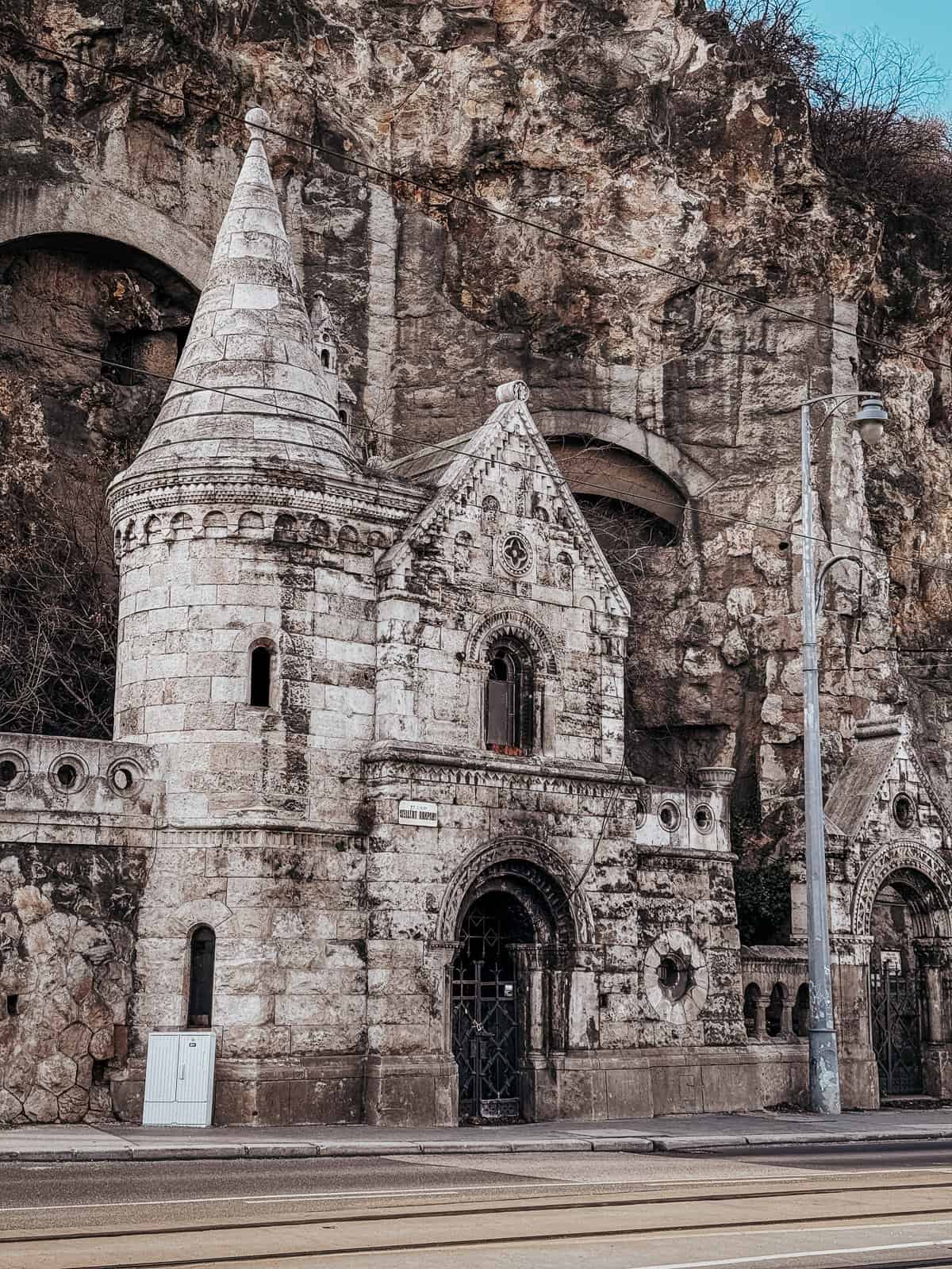 A stone church with a conical roof is built into the rocky hillside, with tram tracks and a lamppost in the foreground.