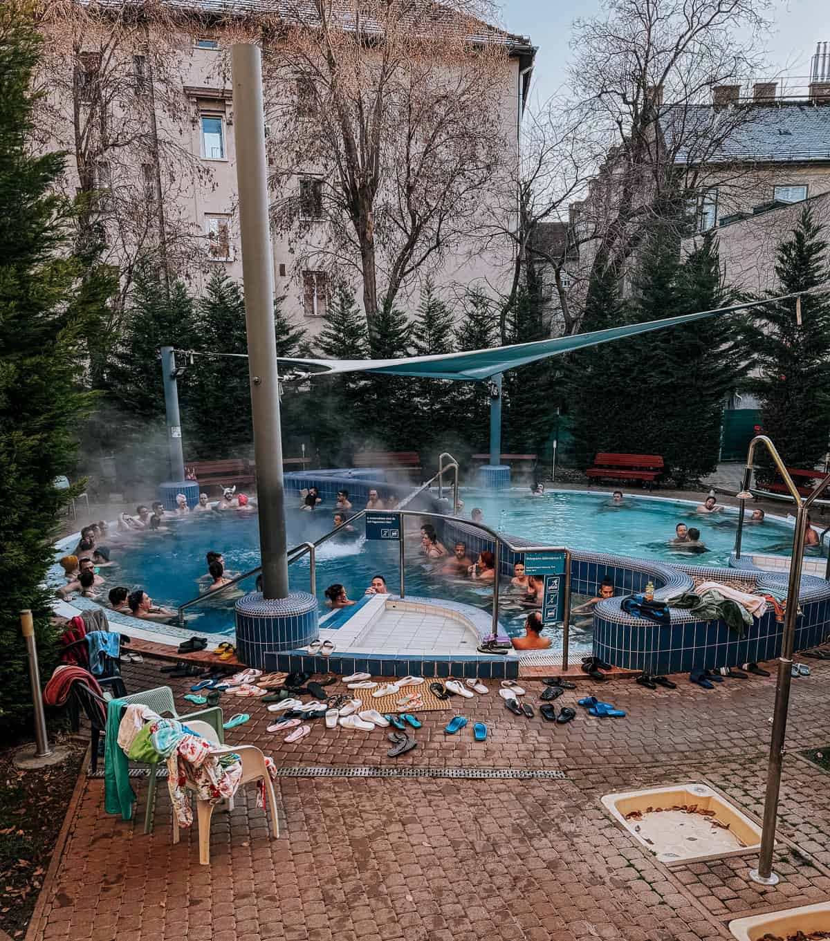 A view of an outdoor thermal bath at Dandar Baths, with people enjoying the hot spring water surrounded by tall trees and apartment buildings. Various sandals and towels are placed around the pool area.