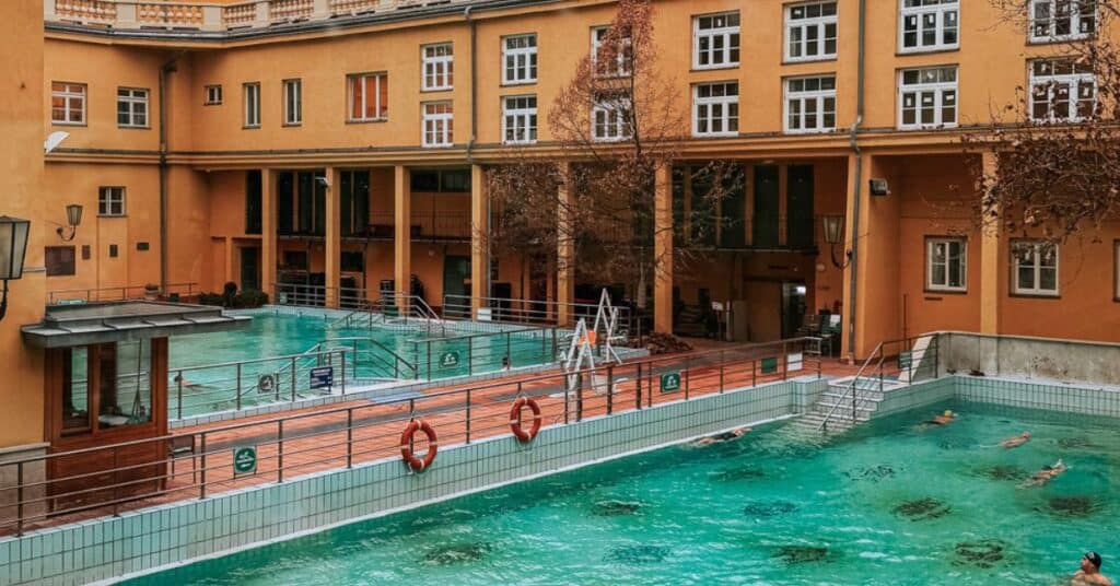 Outdoor pools at Lukács Thermal Bath with clear blue water, surrounded by yellow buildings and lifebuoys along the edges.