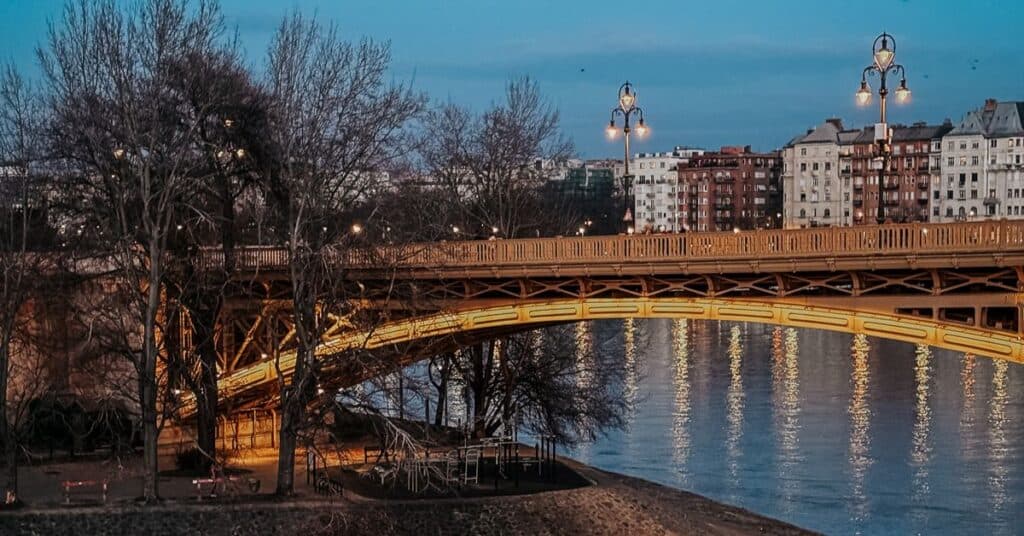 Margaret Island bridge illuminated with warm lights at dusk, reflecting in the river below with buildings in the background on Margaret Island.