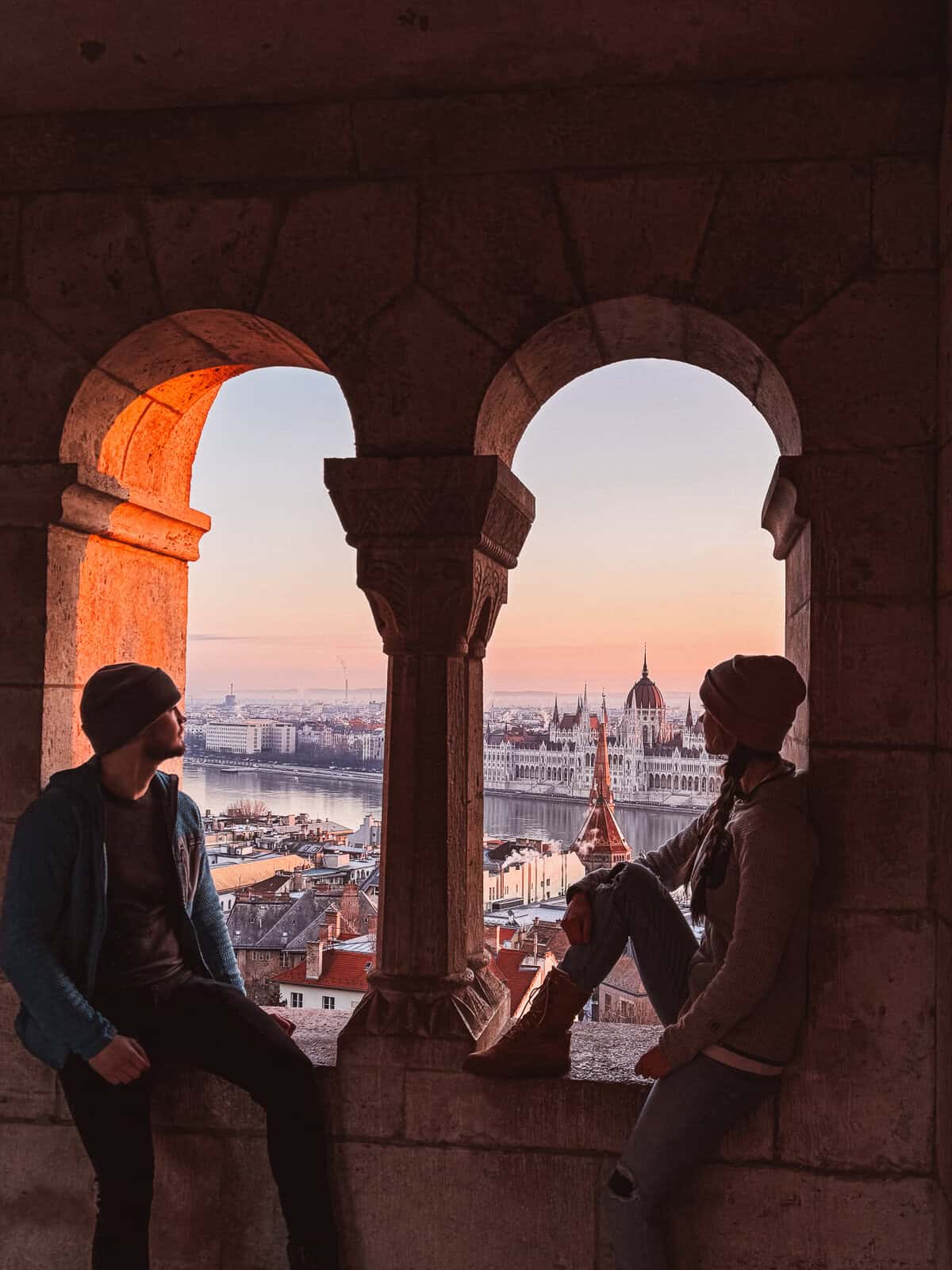 Two people sit within an arched window of the Fisherman’s Bastion, overlooking the Parliament building at sunset.