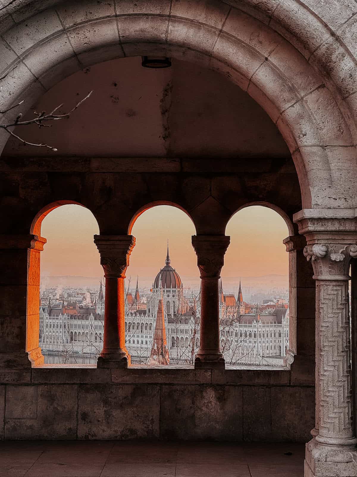 The Hungarian Parliament building viewed through three arches of the Fisherman’s Bastion at sunset, casting an orange glow.