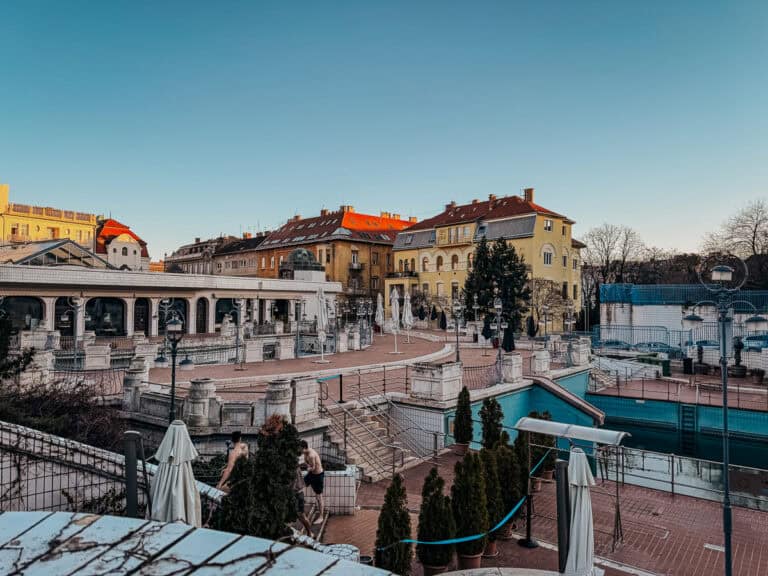 The exterior of Gellert Baths showcasing its historic architecture, with a wide-open courtyard, archways, and decorative elements. The surrounding buildings have a mix of classical and modern styles under a clear blue sky.