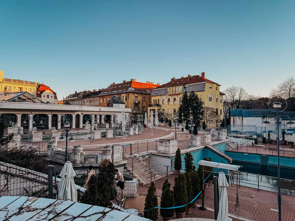 The exterior of Gellert Baths showcasing its historic architecture, with a wide-open courtyard, archways, and decorative elements. The surrounding buildings have a mix of classical and modern styles under a clear blue sky.