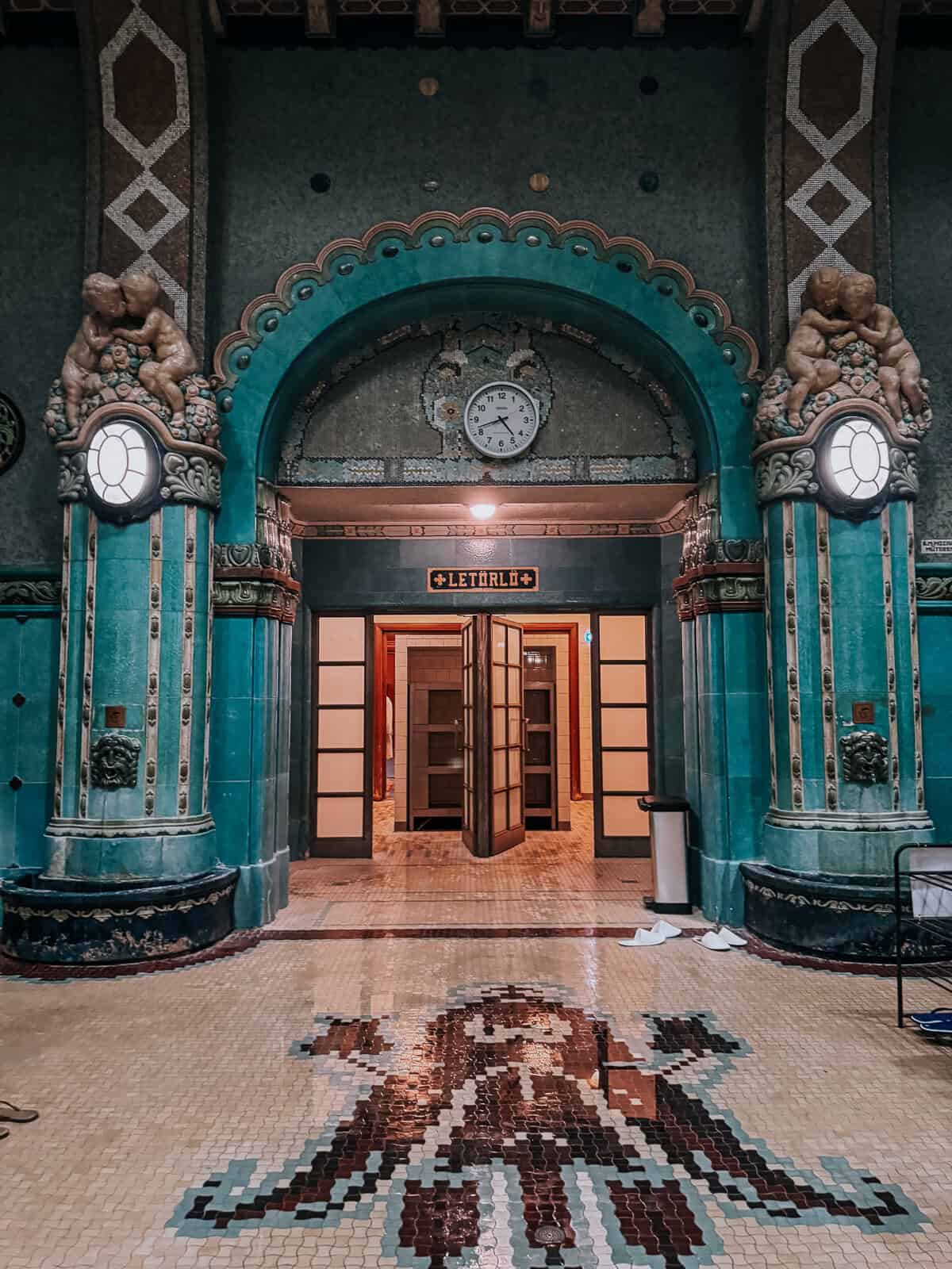 Interior of Gellert Baths featuring a grand entrance with ornate green columns, a mosaic floor, and an arched doorway. The space exudes historical charm with intricate details and a clock above the entrance.