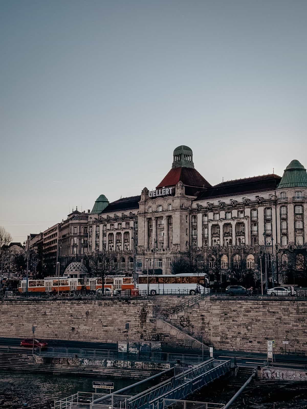 A distant view of the Gellert Baths building, displaying its prominent dome and intricate facade. The building is set against a clear sky, with tram tracks and vehicles visible in front.