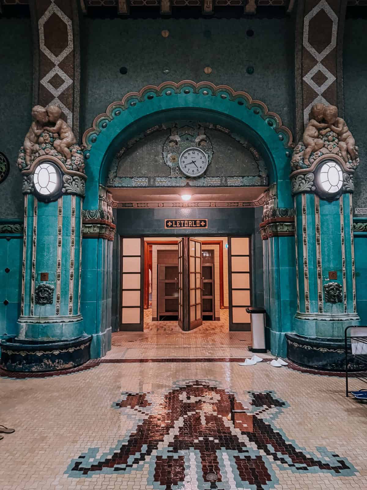 Entrance hall of the Gellért Thermal Bath with intricate mosaic flooring and an arched doorway flanked by decorative columns.