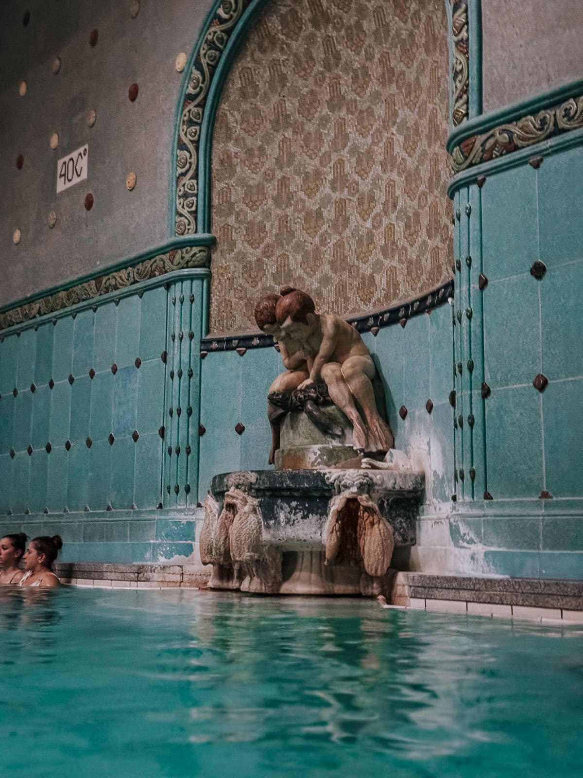 Water fountain in the Gellért Thermal Bath, featuring a statue of a woman and child against a turquoise mosaic wall, with water flowing into the pool below.