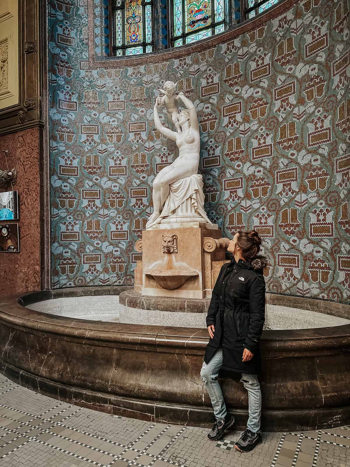 Statue of a woman and child in the Gellért Thermal Bath, set against a backdrop of ornate mosaic tiles.