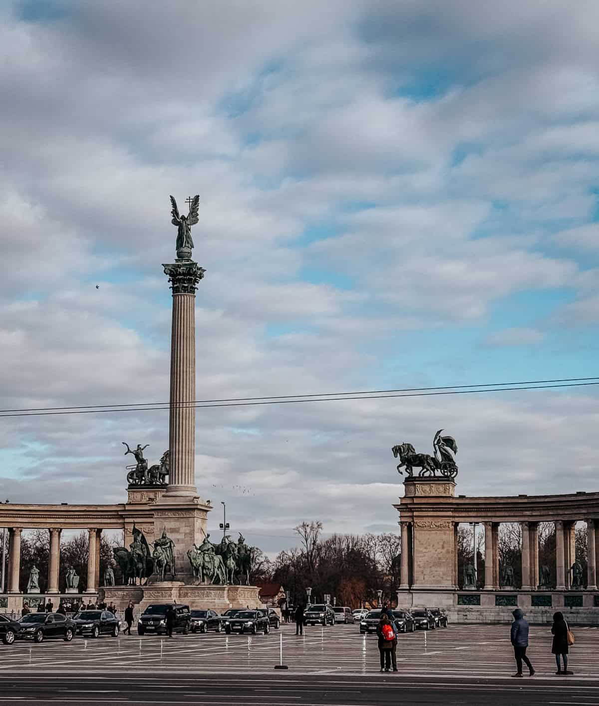 The central part of Heroes' Square showcasing the tall column topped with a statue of Archangel Gabriel, surrounded by other statues and visitors.