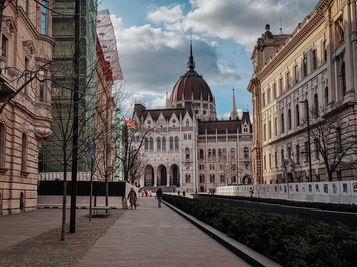 The Hungarian Parliament building viewed from a distance, framed by modern and historical buildings on a walkway.