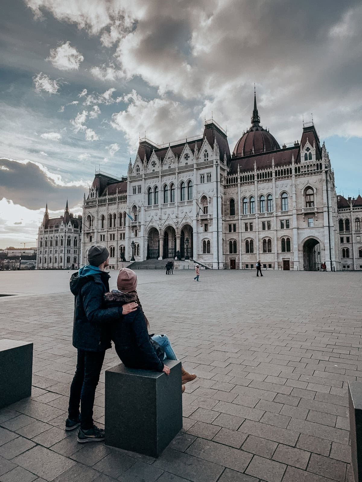 Two people sitting and standing close to each other in front of the Hungarian Parliament building, with a dramatic sky above.