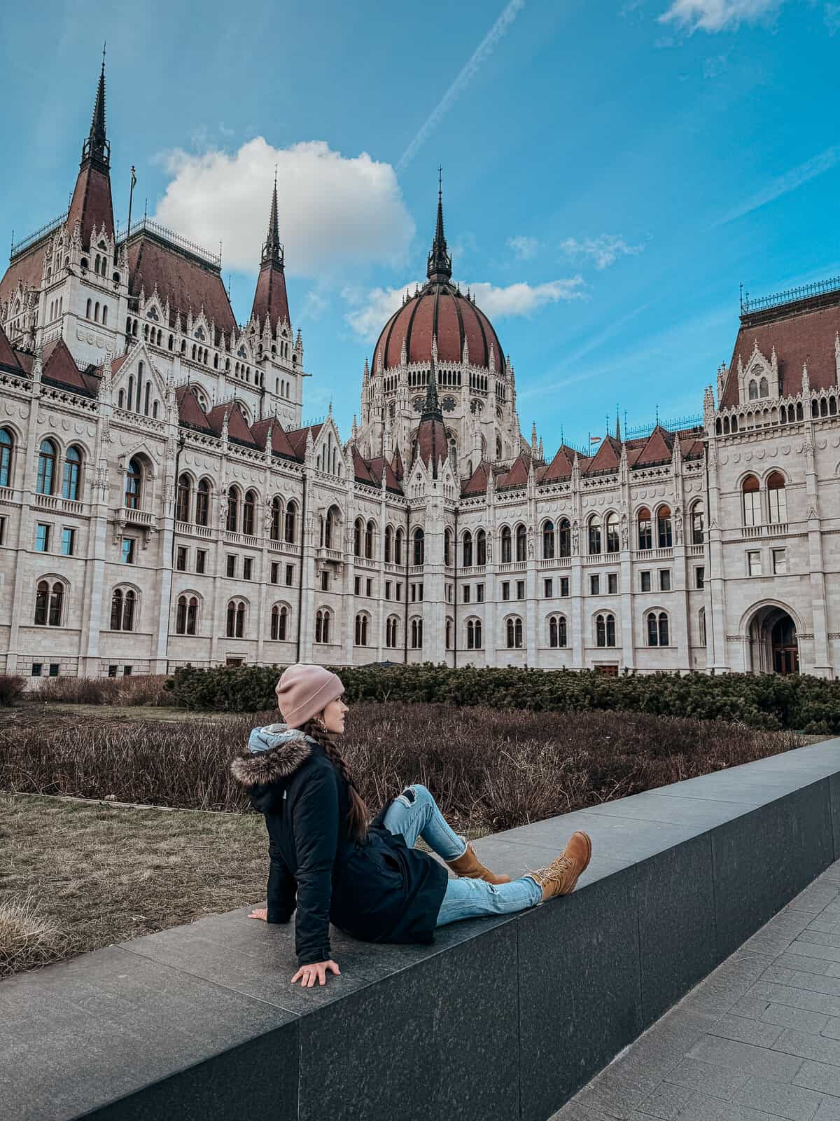 A person sitting on a ledge near the Hungarian Parliament building, enjoying a clear, sunny day.