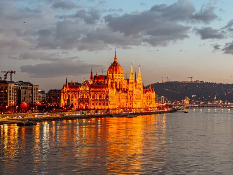 The Hungarian Parliament Building illuminated at dusk, reflecting off the Danube River with a soft orange glow, under a partly cloudy sky.