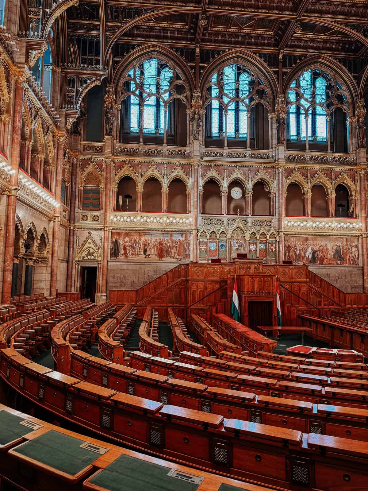 The grand assembly hall of the Hungarian Parliament Building, featuring wooden seats, ornate walls with murals, and large stained glass windows.