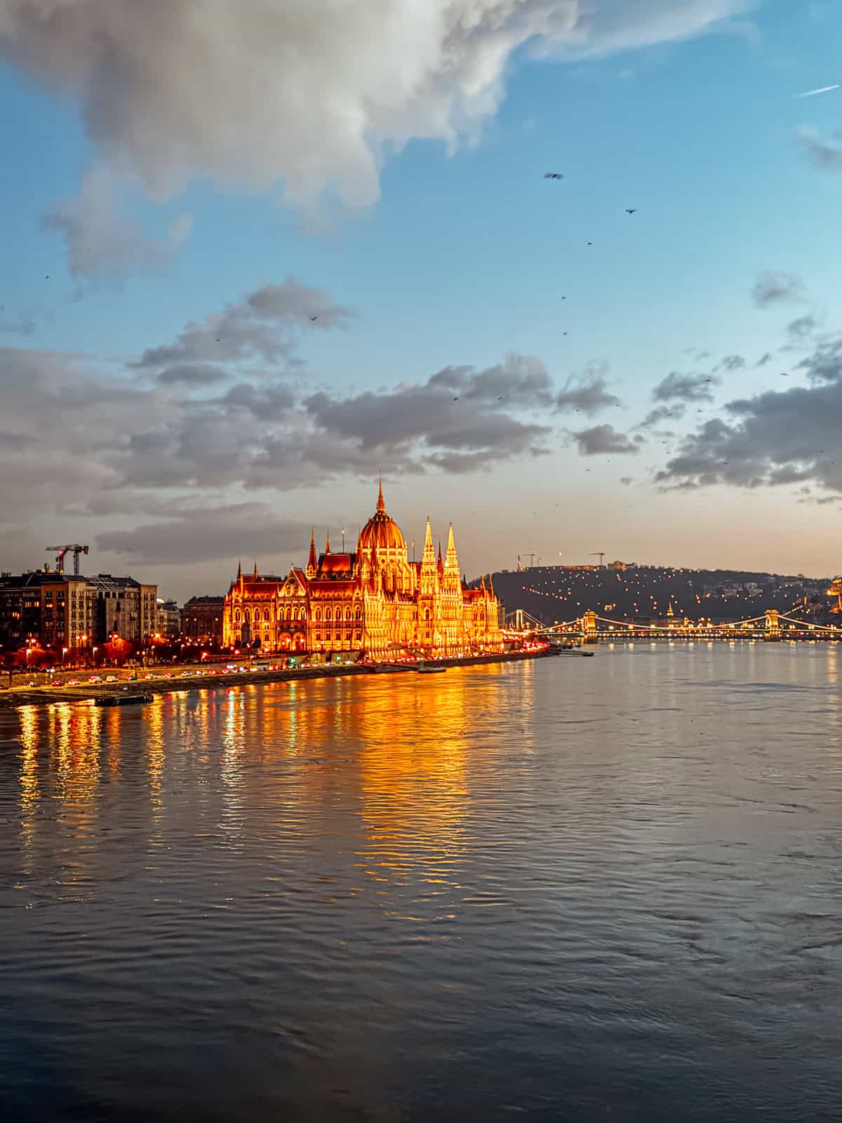 A wider view of the Hungarian Parliament Building at dusk, brightly lit and reflecting in the Danube River, with a serene evening sky in the background.