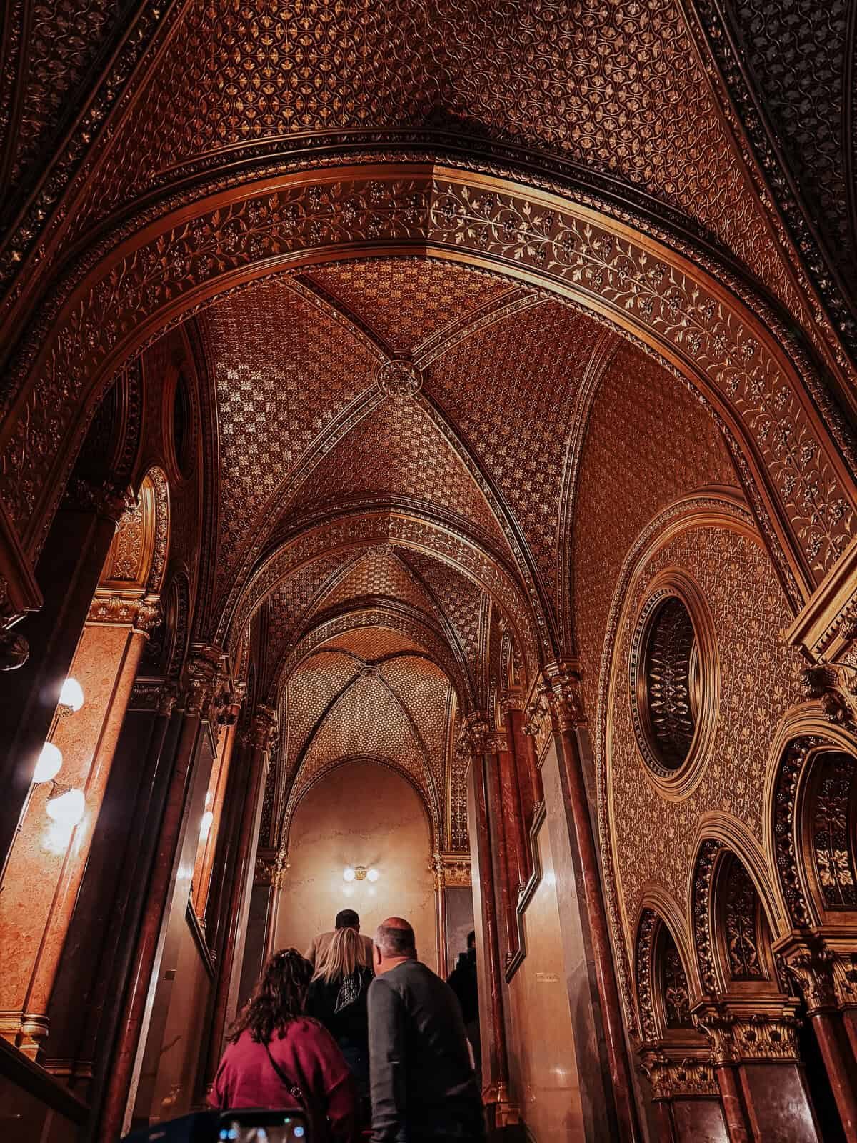 An intricately detailed, golden-colored interior hallway of the Hungarian Parliament Building, featuring arched ceilings and ornate designs.