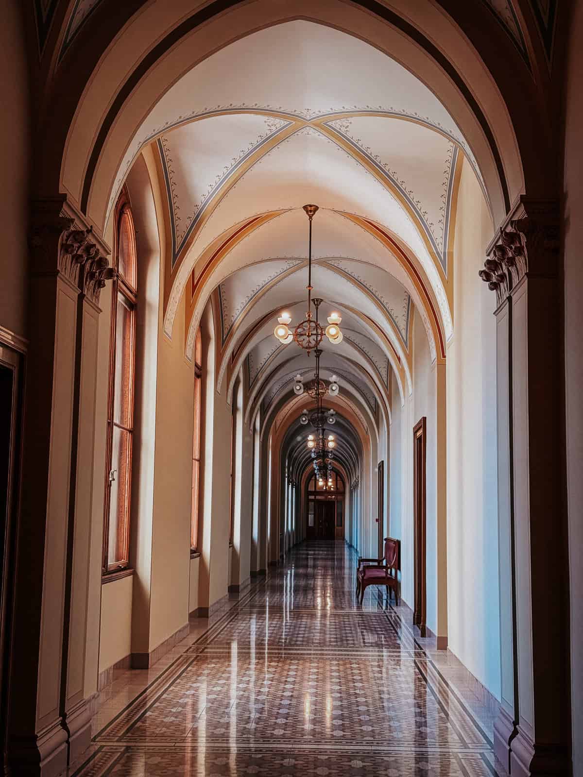 A long, elegant hallway inside the Hungarian Parliament Building, with high arched ceilings, chandeliers, and a checkered tile floor.