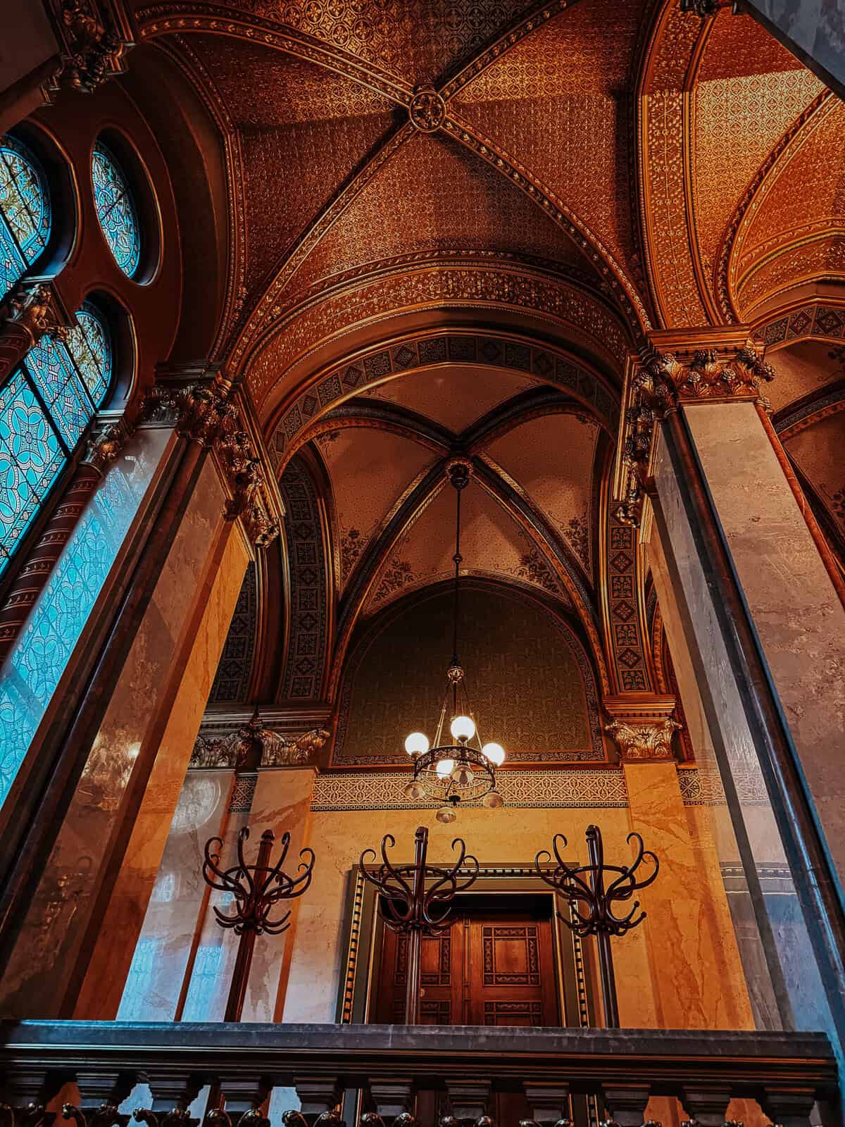 A richly decorated interior section of the Hungarian Parliament Building, showcasing ornate gold arches, detailed ceiling patterns, and large stained glass windows.