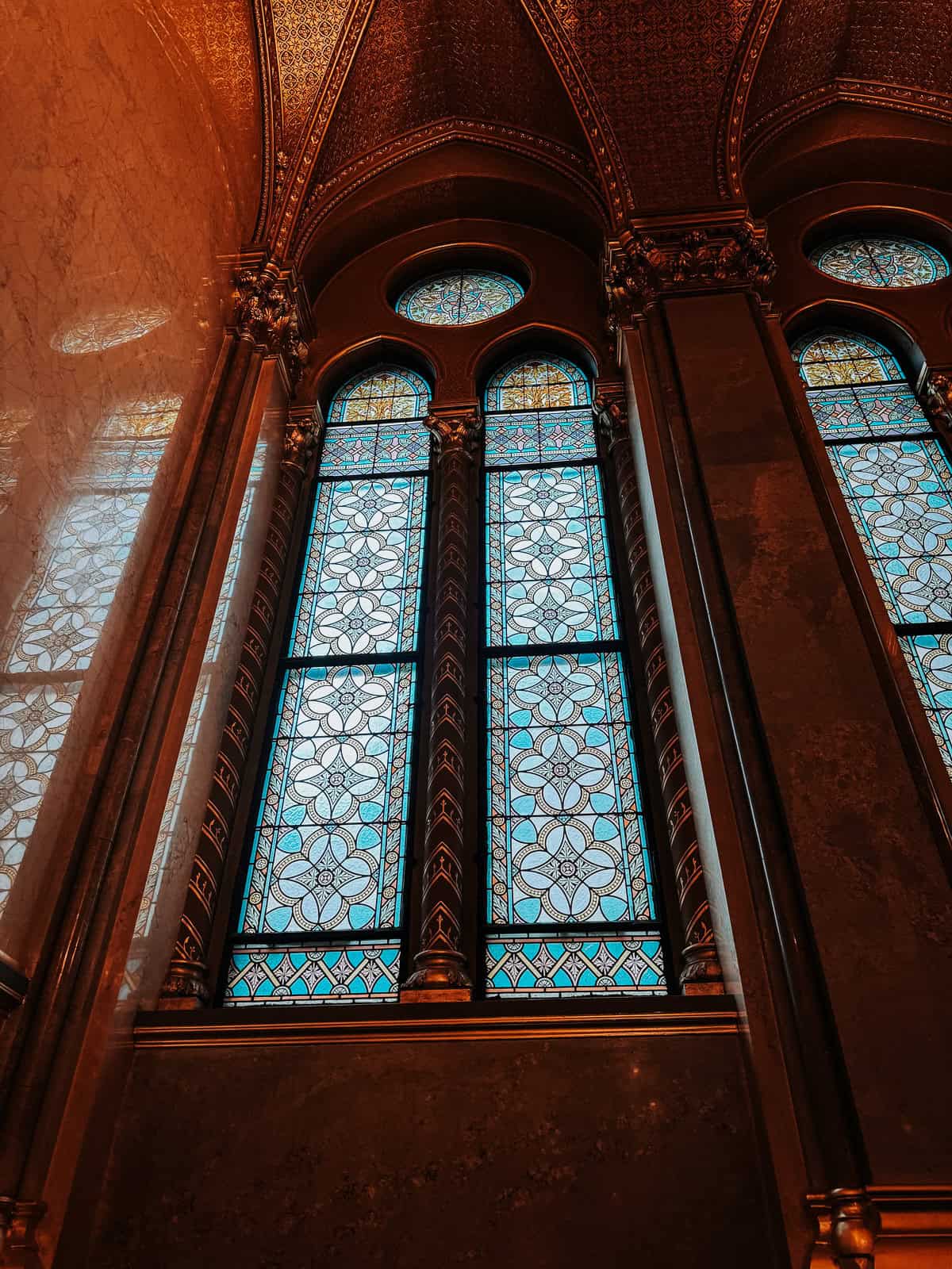 Close-up view of three large, arched stained glass windows within the Hungarian Parliament Building, framed by intricate golden designs.