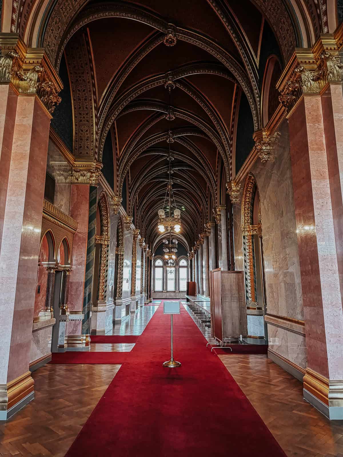 A grand hallway inside the Hungarian Parliament Building with a red carpet, high arches, and luxurious gold detailing on the walls and ceiling.