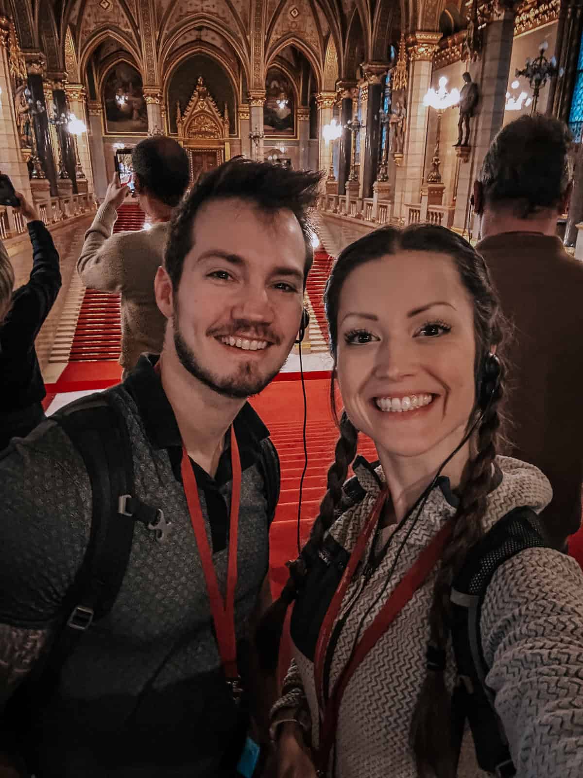 A smiling couple taking a selfie inside the Hungarian Parliament Building, with a background of a richly decorated hall featuring statues, red carpet, and ornate lighting.