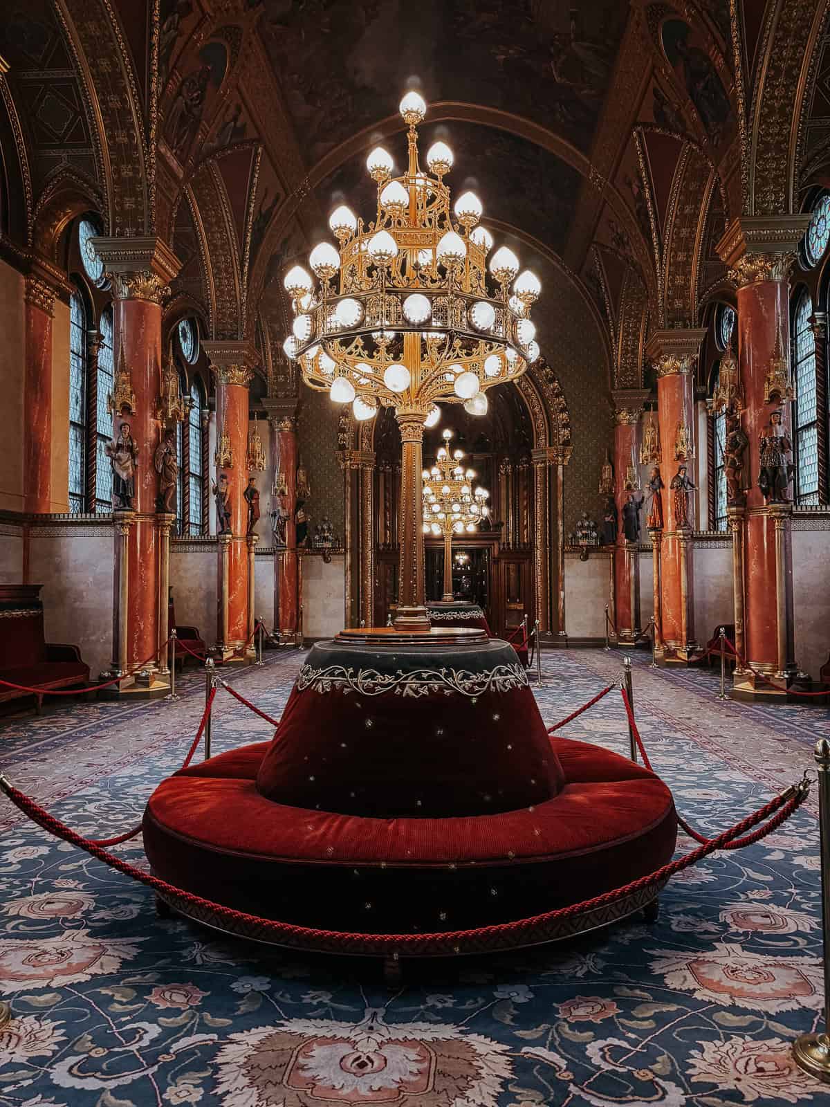 An opulent room in the Hungarian Parliament Building with large, golden chandeliers, red velvet seating, and intricately designed pillars.