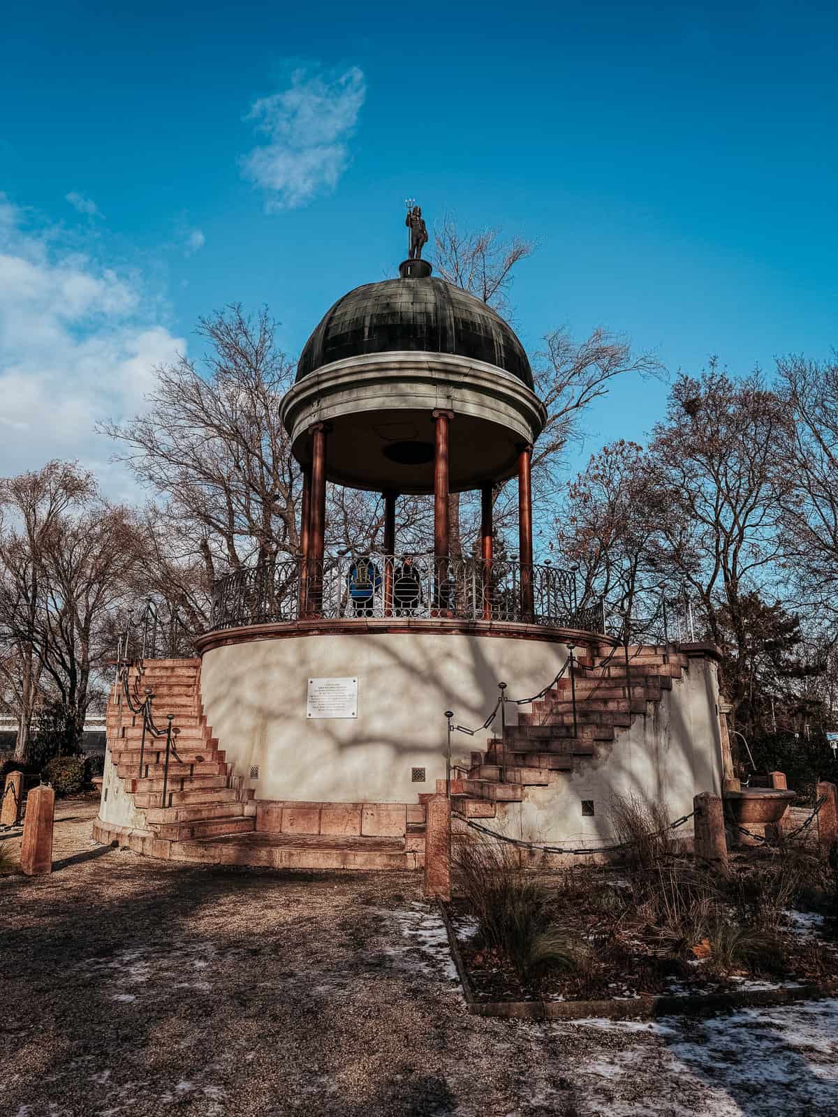 A large, domed gazebo with columns and a staircase in a garden setting, surrounded by trees and under a bright blue sky