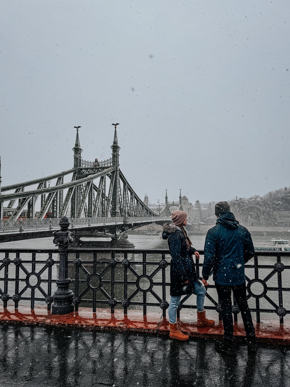 Two people stand by the railing in front of Liberty Bridge in Budapest during a snowfall.