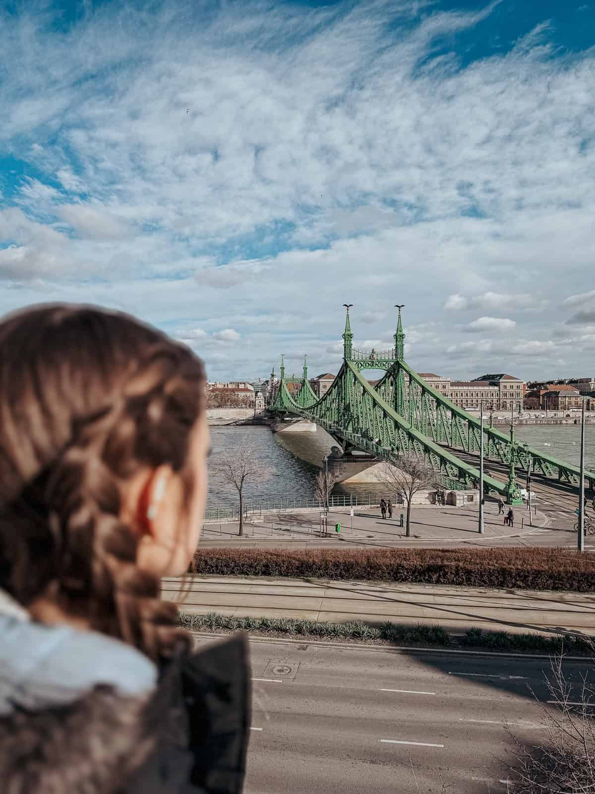 A person with braided hair looks towards Liberty Bridge on a partly cloudy day.