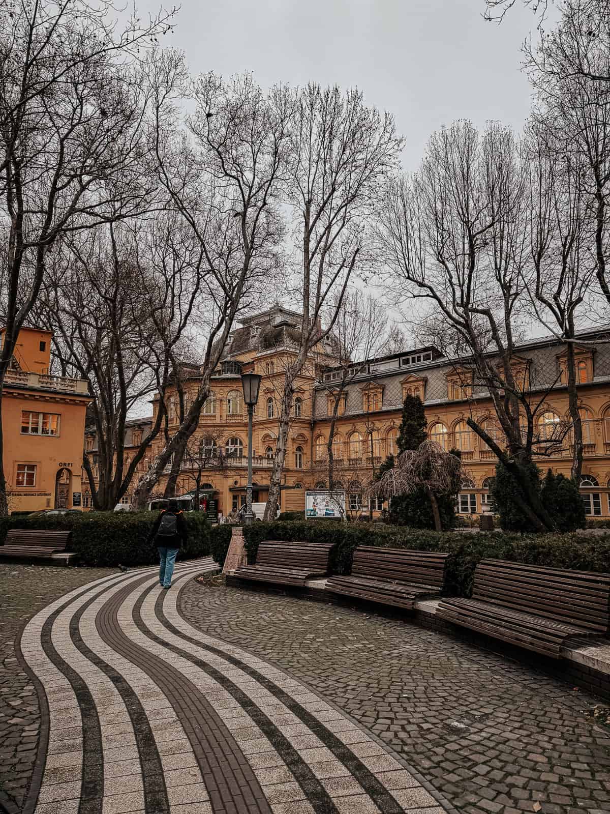 The exterior of Lukacs Baths, featuring a serene courtyard with a winding pathway, benches, and leafless trees. A person is seen walking towards the historic bathhouse building.