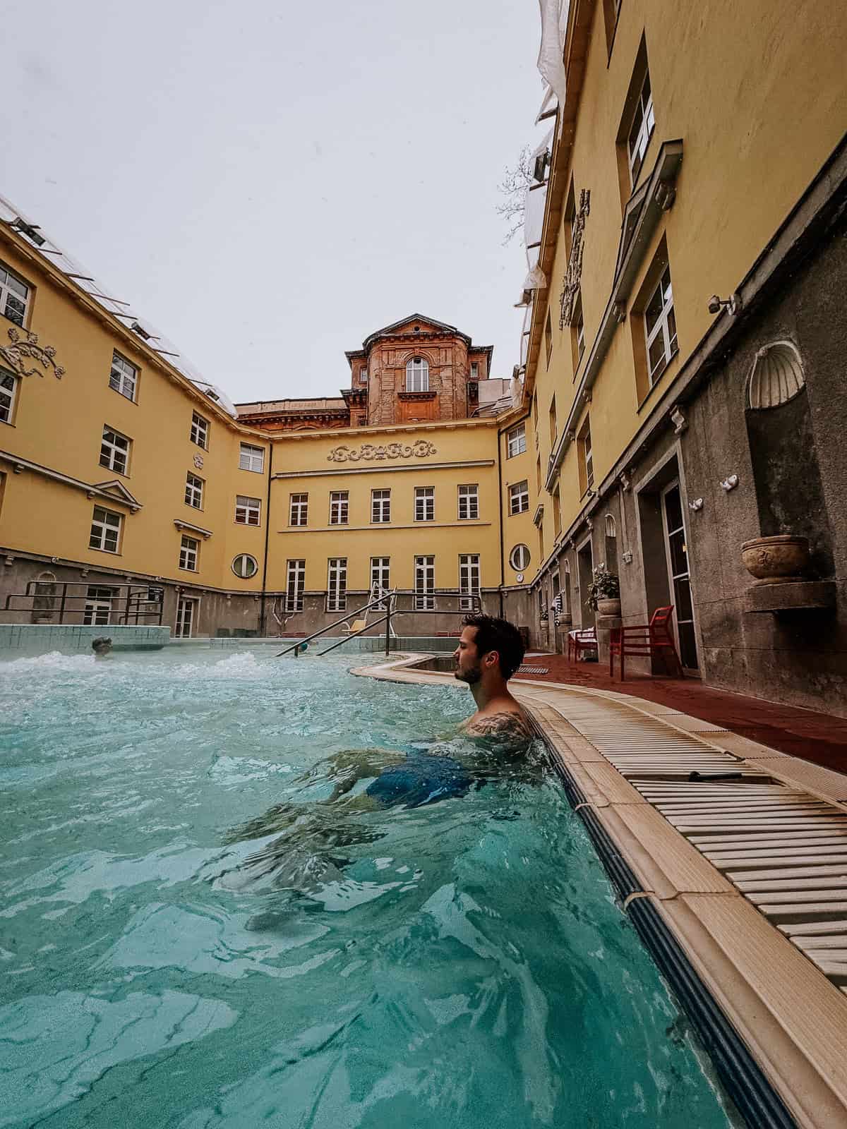 An outdoor pool at Lukacs Baths, with a man relaxing in the warm water. The surrounding yellow buildings and open sky create a calm and inviting atmosphere.