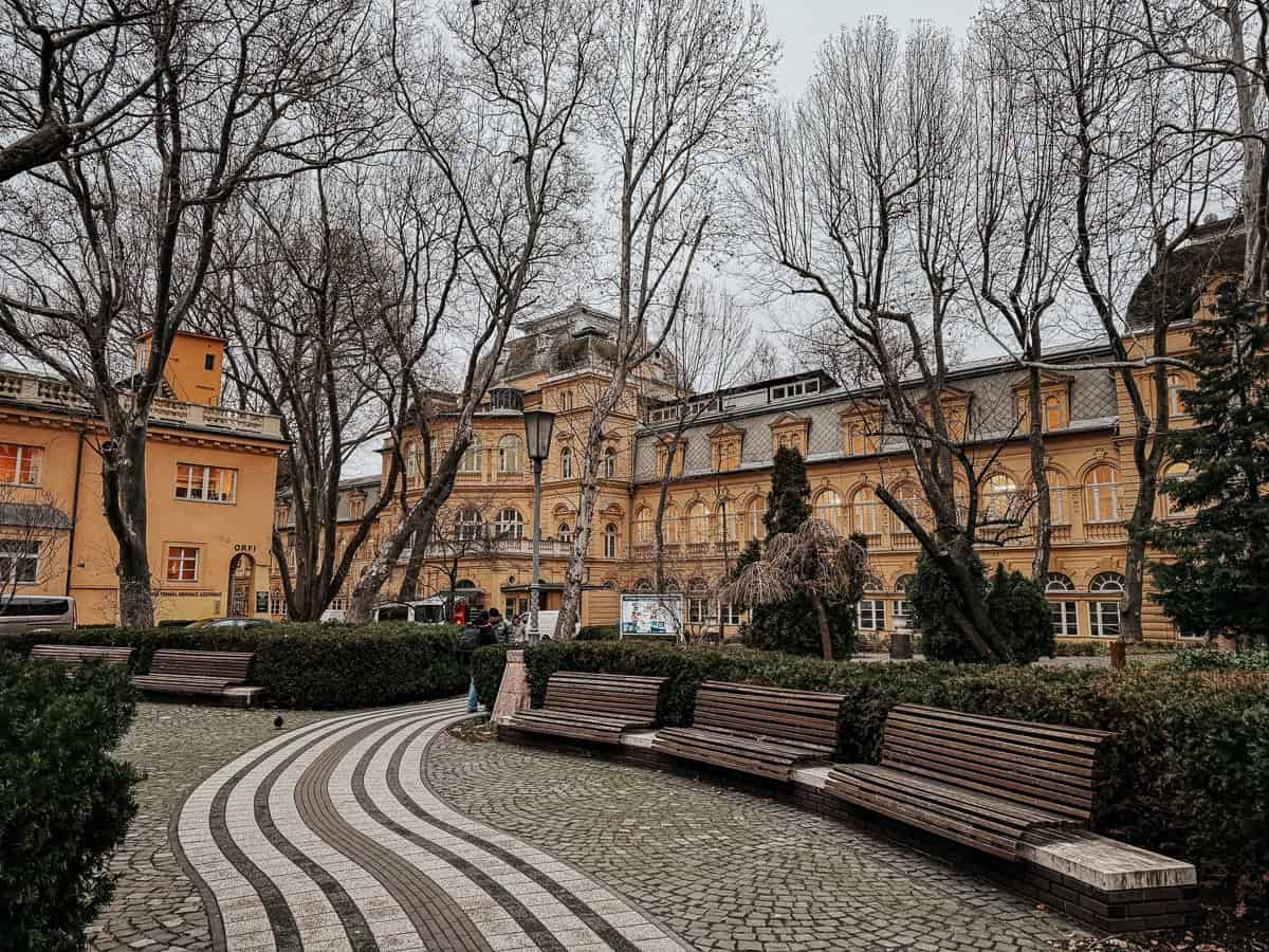 A scenic view of the exterior of Lukács Thermal Bath, showing historic yellow buildings surrounded by leafless trees in a park setting with curving pathways and benches.